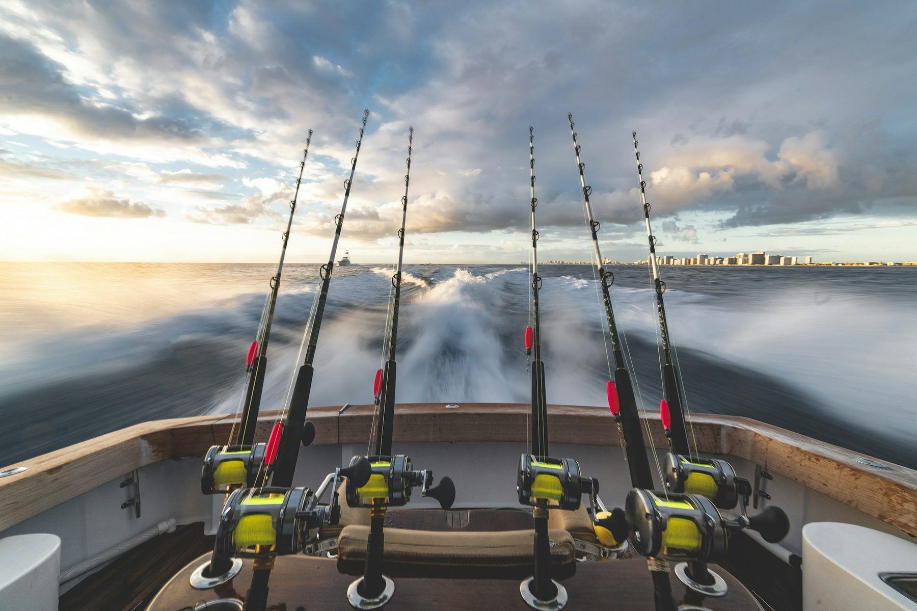 Six heavy trolling rods with gold reels in a sportfishing boat's cockpit, wake churning under a dramatic sky